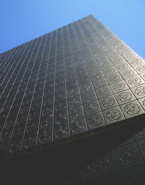 A modern building facade covered with dark metal panels featuring intricate geometric patterns. The structure extends upwards against a clear blue sky, creating a sense of height and modern architectural design.