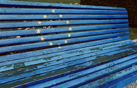 A blue wooden bench features the European Union flag design, with yellow stars arranged in a circular pattern against the blue slats. The paint appears slightly worn, giving the bench a weathered look. The bench is placed on a concrete surface with greenery visible in the background.
