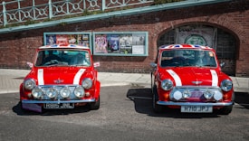 Two classic red Mini cars are parked side by side against a backdrop of a brick wall adorned with posters. Both cars feature white racing stripes and Union Jack designs on their roofs and side mirrors. They are parked on a paved area with a metal fence on top of the wall, and various advertisements are displayed on the wall.
