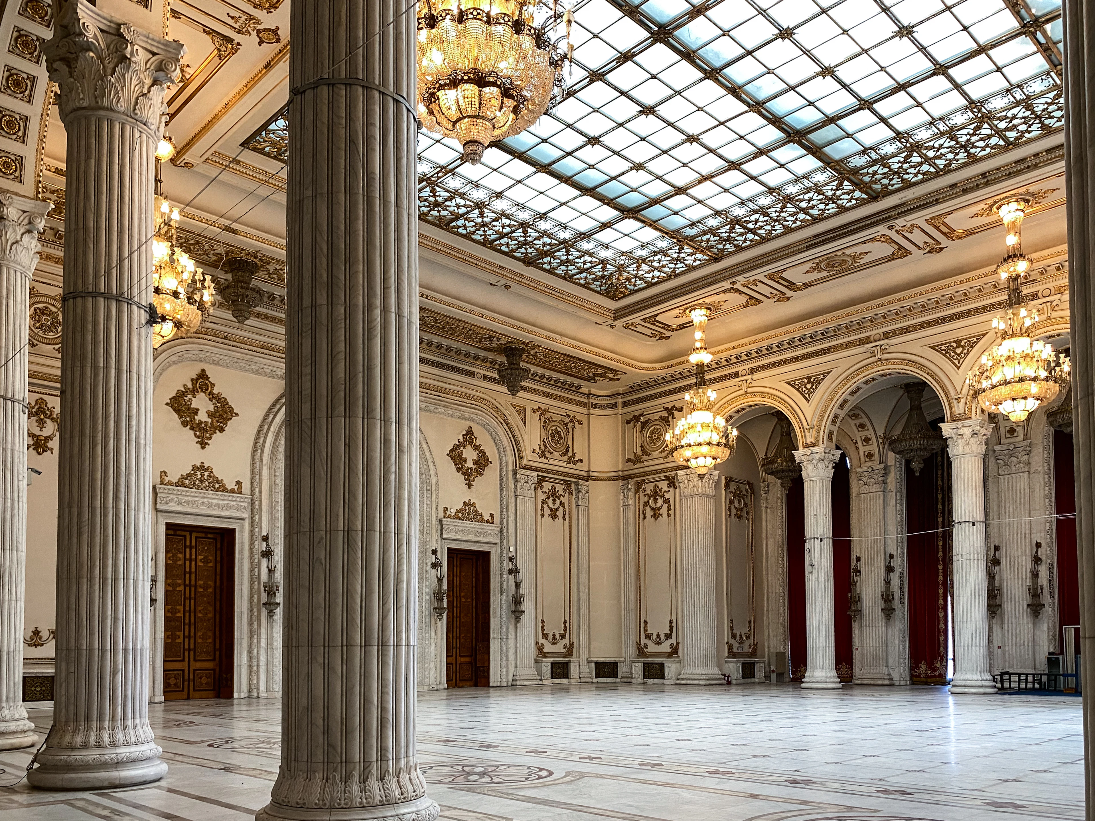 Marble columns and crystal chandeliers fill a grand hall inside the Palace of Parliament beneath a glass ceiling.
