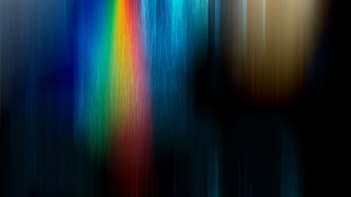Vertical streaks of color blend together, creating a visually striking pattern. A vibrant rainbow-like gradient transitions from blue to green, yellow, and red against a dark and blurred background, evoking a sense of motion or digital abstraction.
