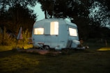 A small vintage camper is situated in a grassy area surrounded by trees. The interior of the camper emits a warm, inviting light through the windows. An outdoor setup includes a chair, a small table, and an umbrella to the left. The scene is set during dusk or early evening, creating a cozy atmosphere.
