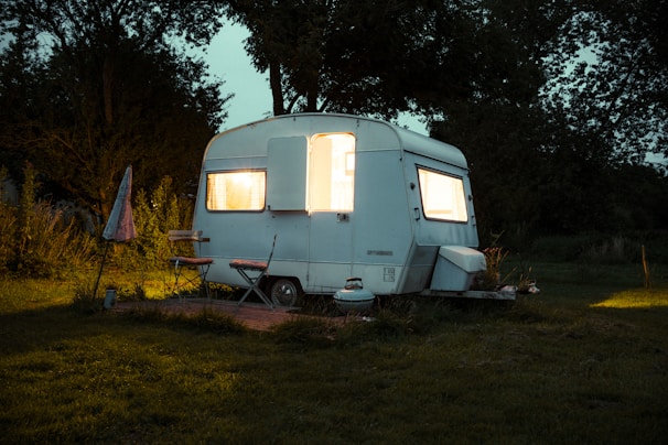 A small vintage camper is situated in a grassy area surrounded by trees. The interior of the camper emits a warm, inviting light through the windows. An outdoor setup includes a chair, a small table, and an umbrella to the left. The scene is set during dusk or early evening, creating a cozy atmosphere.