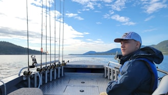 A person wearing a cap and jacket is seated on a metal boat deck, facing forward into a scenic view of water, mountains, and a partly cloudy sky. Multiple fishing rods are lined up at the side of the boat.