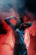 A person is standing amidst swirling, colorful smoke. The lighting casts dramatic shadows, and the outfit reflects a sheen, giving a futuristic or ethereal appearance.