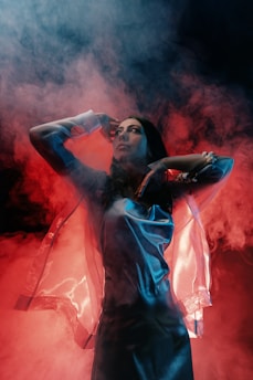 A person is standing amidst swirling, colorful smoke. The lighting casts dramatic shadows, and the outfit reflects a sheen, giving a futuristic or ethereal appearance.