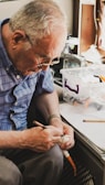 An elderly person with gray hair and wearing glasses is intently focused on an activity. They are holding what appears to be a carving tool in one hand and a small object in the other, possibly engaged in a craft or hobby. A transparent plastic box with materials and tools is on the table nearby.