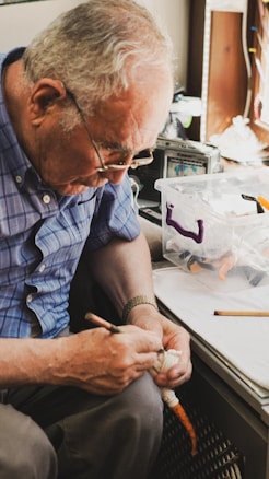 An elderly person with gray hair and wearing glasses is intently focused on an activity. They are holding what appears to be a carving tool in one hand and a small object in the other, possibly engaged in a craft or hobby. A transparent plastic box with materials and tools is on the table nearby.