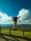 A person wearing casual outdoor clothing stands on a wooden fence with arms outstretched, looking at a vast, scenic landscape of green hills under a bright blue sky filled with fluffy white clouds.