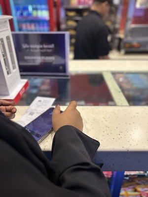 A person in a black attire is standing at a counter, holding a smartphone and a receipt. Behind the counter, a worker in black uniform is partially visible, with a background of shelves and machines, possibly in a store or a café. Nearby, a sign in two languages is displayed, and there are packaged products on the shelves.