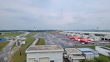 A modern airport scene with several aircraft parked near a terminal. The planes are branded with a prominent airline logo, and the air traffic control tower is visible in the distance. The infrastructure includes runways, roads, and buildings, surrounded by grassy areas under a cloudy sky.