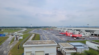 A modern airport scene with several aircraft parked near a terminal. The planes are branded with a prominent airline logo, and the air traffic control tower is visible in the distance. The infrastructure includes runways, roads, and buildings, surrounded by grassy areas under a cloudy sky.