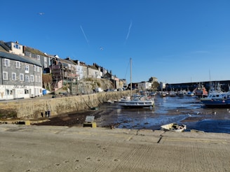 A coastal town harbor with several boats docked in low tide, revealing the muddy ground. The background shows a row of traditional buildings with a café named 'She Sells'. The sky is clear with a few seagulls flying overhead.