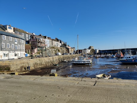 A coastal town harbor with several boats docked in low tide, revealing the muddy ground. The background shows a row of traditional buildings with a café named 'She Sells'. The sky is clear with a few seagulls flying overhead.