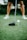 A close-up view of a golf club poised next to a white golf ball on a tee, with a player’s legs visible in the background standing on a lush green golf course.