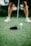 A close-up view of a golf club poised next to a white golf ball on a tee, with a player’s legs visible in the background standing on a lush green golf course.