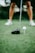 A close-up view of a golf club poised next to a white golf ball on a tee, with a player’s legs visible in the background standing on a lush green golf course.