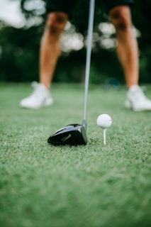 A close-up view of a golf club poised next to a white golf ball on a tee, with a player&rsquo;s legs visible in the background standing on a lush green golf course.