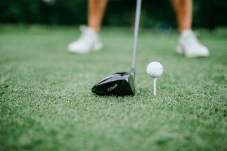 A close-up of a golf club positioned next to a golf ball on a tee. The focus is on the club head and the ball, with blurred legs wearing white shoes in the background.