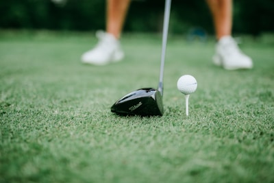 A close-up of a golf club positioned next to a golf ball on a tee. The focus is on the club head and the ball, with blurred legs wearing white shoes in the background.