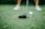 A close-up of a golf club positioned next to a golf ball on a tee. The focus is on the club head and the ball, with blurred legs wearing white shoes in the background.