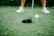 A close-up of a golf club positioned next to a golf ball on a tee. The focus is on the club head and the ball, with blurred legs wearing white shoes in the background.