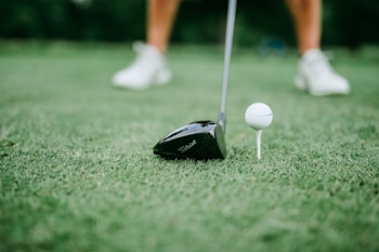 A close-up of a golf club positioned next to a golf ball on a tee. The focus is on the club head and the ball, with blurred legs wearing white shoes in the background.