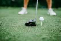 A close-up of a golf club positioned next to a golf ball on a tee. The focus is on the club head and the ball, with blurred legs wearing white shoes in the background.