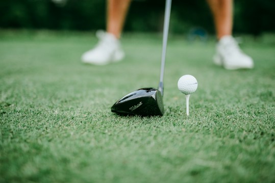 A close-up of a golf club positioned next to a golf ball on a tee. The focus is on the club head and the ball, with blurred legs wearing white shoes in the background.