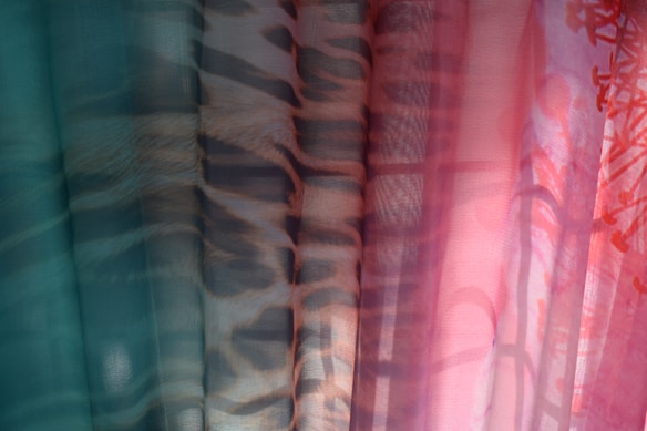 A series of sheer fabric panels with varying patterns and colors, creating a layered effect. The fabrics include teal, animal print, and floral designs with a soft light filtering through, enhancing the textures and colors.