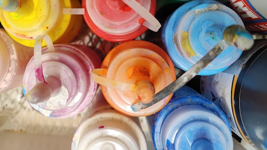 A collection of colorful squeeze bottles arranged in a circular pattern, viewed from above. Each bottle features a distinct color, including yellow, red, orange, blue, white, and purple, with slight paint splatters visible on their surfaces. The bottles are packed closely together, and some appear to show signs of use with dried paint on their tips.