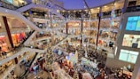 A bustling, multi-level shopping mall decorated for the holiday season, featuring a large Christmas tree at the center surrounded by people shopping and walking. Bright lights and festive decorations are hung throughout the balconies and escalators. The area is crowded with shoppers, and various stores are visible on multiple floors.
