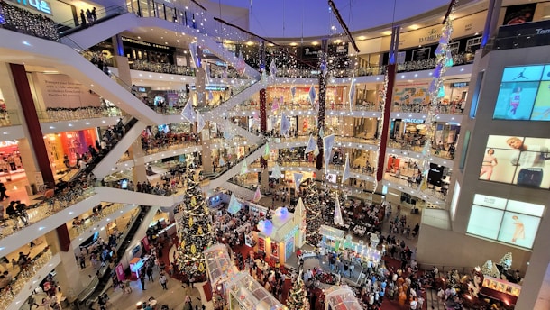 A bustling, multi-level shopping mall decorated for the holiday season, featuring a large Christmas tree at the center surrounded by people shopping and walking. Bright lights and festive decorations are hung throughout the balconies and escalators. The area is crowded with shoppers, and various stores are visible on multiple floors.