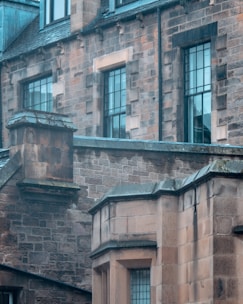 An architectural detail of a historic stone building featuring large rectangular windows and intricate stonework. The facade is made of brown and grey bricks, with visible weathering and moss indicating age. The overall style suggests a traditional or Gothic influence.
