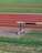 An athletic track with multiple lanes marked in red and green, featuring grass areas alongside. A hurdle is placed near the track on a paved surface.