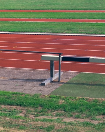 An athletic track with multiple lanes marked in red and green, featuring grass areas alongside. A hurdle is placed near the track on a paved surface.