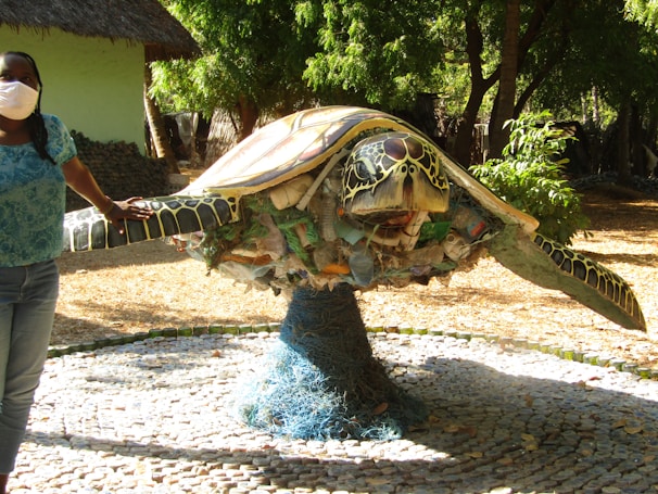 A large sculpture of a turtle made from various pieces of plastic and debris is displayed outdoors on a platform surrounded by trees. A person wearing a mask and a blue shirt stands beside it, placing a hand on the turtle's shell. The sculpture showcases intricate details of the turtle's features and highlights environmental themes.