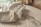 A plush white bathrobe and a leather toiletry bag are placed on a neatly made bed with a branded Costa Verde Express design on the sheets. A window in the background provides soft natural lighting.