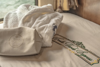 A plush white bathrobe and a leather toiletry bag are placed on a neatly made bed with a branded Costa Verde Express design on the sheets. A window in the background provides soft natural lighting.