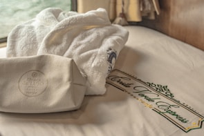 A plush white bathrobe and a leather toiletry bag are placed on a neatly made bed with a branded Costa Verde Express design on the sheets. A window in the background provides soft natural lighting.