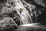 A person wearing a yellow backpack is descending a steep waterfall using ropes. The surrounding rocky landscape is rugged, with water cascading down in a dynamic rush.