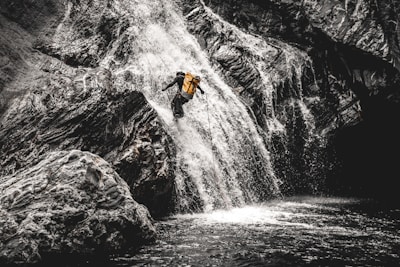A person wearing a yellow backpack is descending a steep waterfall using ropes. The surrounding rocky landscape is rugged, with water cascading down in a dynamic rush.