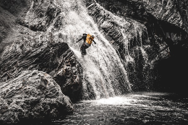 A person wearing a yellow backpack is descending a steep waterfall using ropes. The surrounding rocky landscape is rugged, with water cascading down in a dynamic rush.