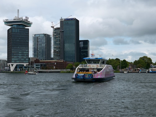 A modern ferry is navigating through a body of water with tall, contemporary buildings in the background. The sky is overcast with clouds, and the ferry appears to be carrying passengers. Green trees and smaller structures can be seen along the shoreline.