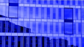 A modern building facade with reflective glass panels creating a grid-like pattern. Vertical lines in the glass alternate between opaque and transparent, allowing partial views of the building's surroundings. The overall blue tint gives the structure an abstract and futuristic look.