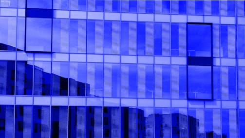 A modern building facade with reflective glass panels creating a grid-like pattern. Vertical lines in the glass alternate between opaque and transparent, allowing partial views of the building's surroundings. The overall blue tint gives the structure an abstract and futuristic look.
