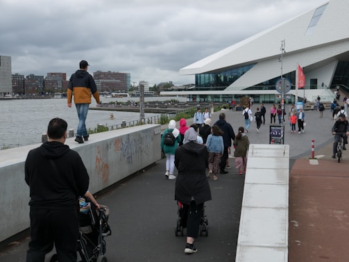 A diverse group of people strolls along a concrete walkway next to a river, with a modern, angular building on the right. The sky is overcast, and there are a few cyclists and people with strollers in the scene.