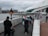 A diverse group of people strolls along a concrete walkway next to a river, with a modern, angular building on the right. The sky is overcast, and there are a few cyclists and people with strollers in the scene.