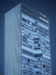 A modern skyscraper with a sleek, glass exterior featuring geometric patterns. The building has a high-rise structure with sharp lines and reflective windows. There is signage in an Asian script at the top.