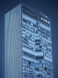 A modern skyscraper with a sleek, glass exterior featuring geometric patterns. The building has a high-rise structure with sharp lines and reflective windows. There is signage in an Asian script at the top.